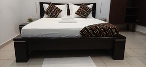 A modern bedroom with a large bed, dark wooden headboard, white bedding, and patterned pillows. - Sheyllo Suites Abidjan (Abidjan)