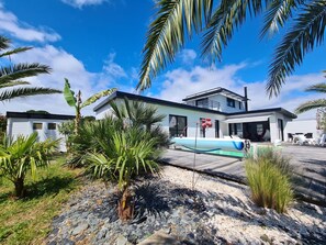 Ein modernes Haus mit Pool, umgeben von tropischen Pflanzen und blauem Himmel.