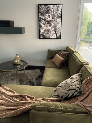 Ein Wohnzimmer mit einem grünen Sofa, einem runden Couchtisch, einem gerahmten Bild an der Wand und Blick durch ein Fenster.