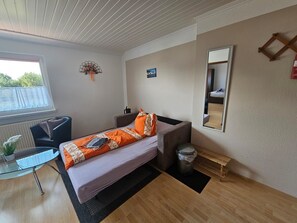 Ein kleines Hotelzimmer mit einem Einzelbett, einem Schreibtisch, einem Stuhl, einem Fenster und einem Spiegel.