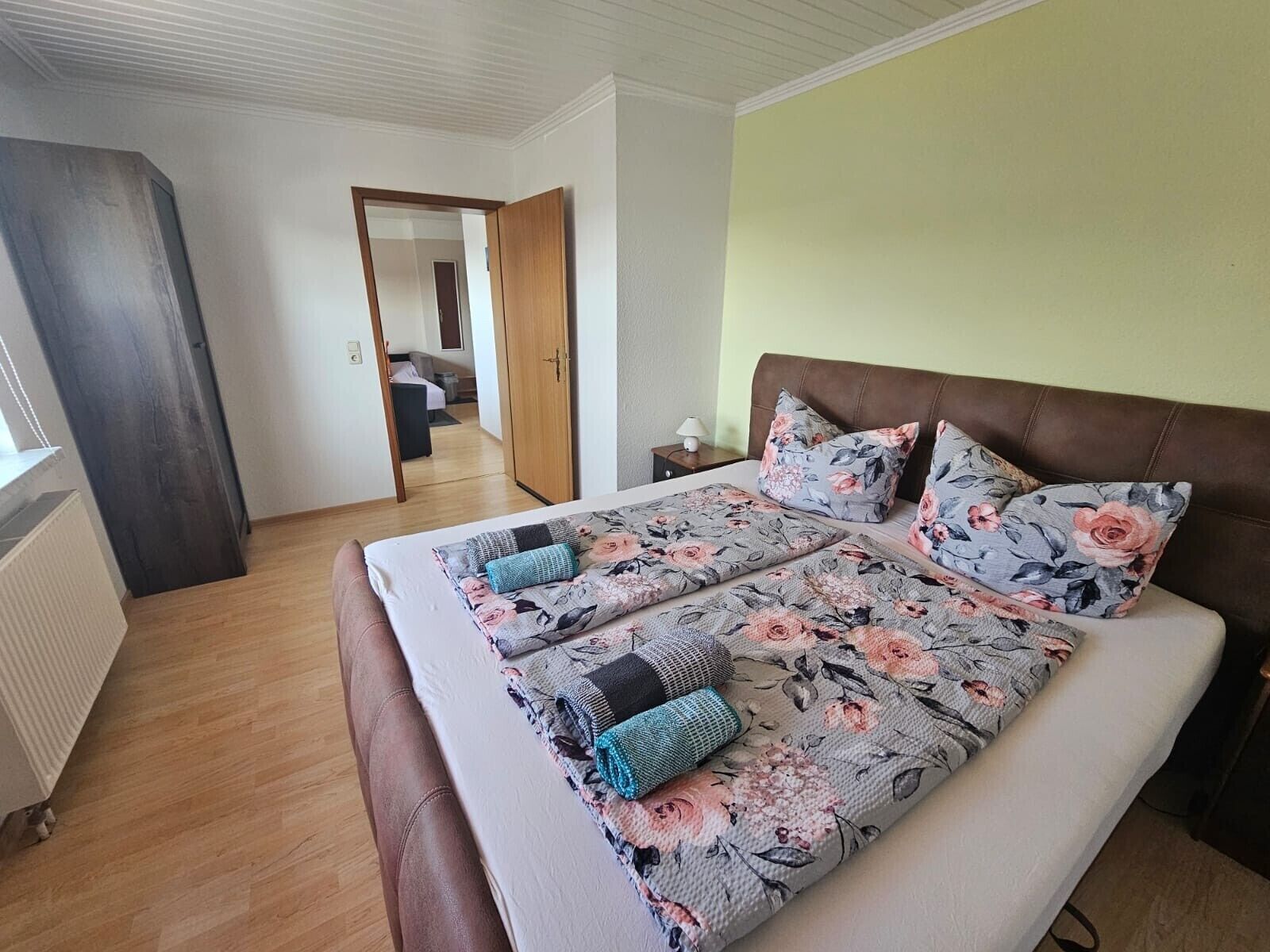 Ein Schlafzimmer mit Bett, Nachttisch, Lampe und Blick in einen anderen Raum durch eine offene Tür.
