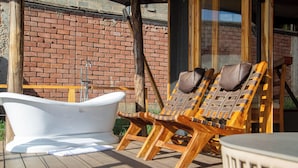 Una terrazza in legno con una vasca da bagno bianca, due poltrone di legno e un muro di mattoni sullo sfondo.