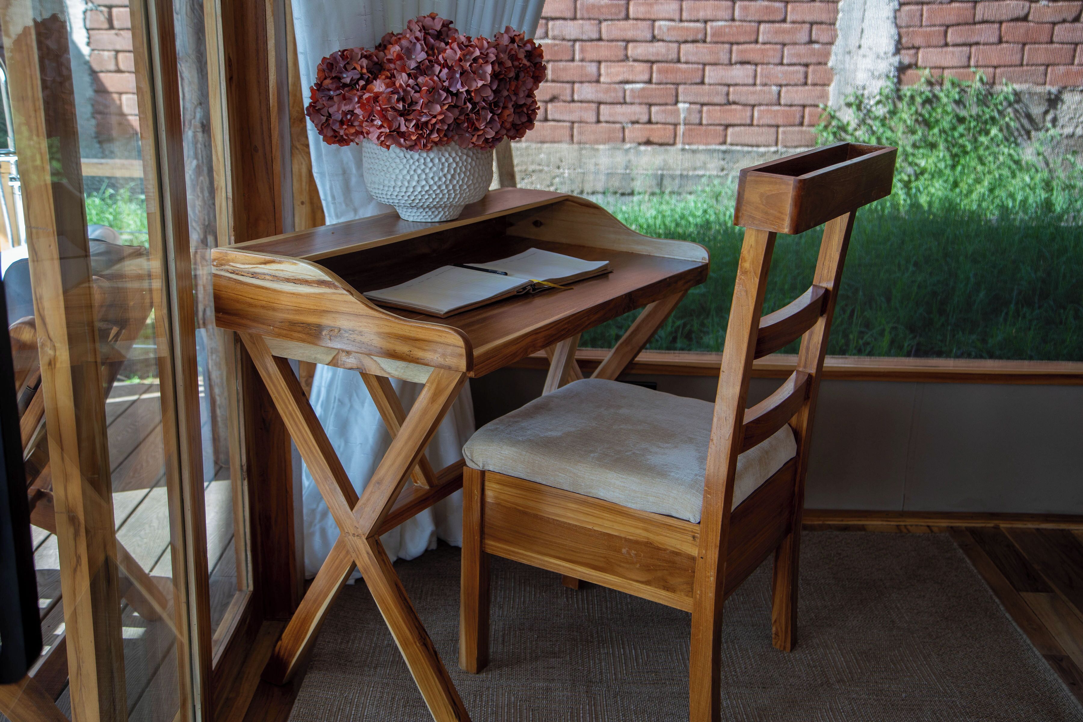 Een houten bureau met een stoel, een vaas met bloemen en een raam met uitzicht op groen.