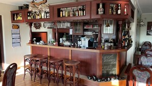 Uma área de bar com banquetas de madeira, um quadro negro com o menu e diversos garrafas e copos.