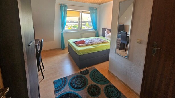 Um quarto com cama, escrivaninha, cadeira, janela com cortinas e teto de madeira.