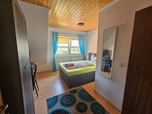 A bedroom with a bed, a desk, a chair, a window with curtains, and a wooden ceiling. - Room 'Pension Dalina - Nr. 17' with Shared Terrace, Shared Garden and Wi-Fi (Sagard)