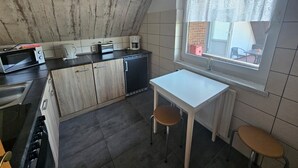 Cocina compacta con microondas, tostadora y cafetera sobre la encimera. Hay una pequeña mesa con dos taburetes y una ventana con vistas al exterior.