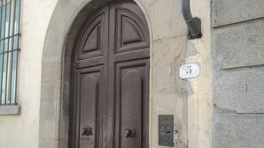 Eine dunkle Holztür mit schlichtem, rundbogigen Eingang, einem kleinen, vergitterten Fenster und einem Schild mit der Hausnummer an der Wand.