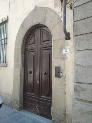 Eine dunkle Holztür mit schlichtem, rundbogigen Eingang, einem kleinen, vergitterten Fenster und einem Schild mit der Hausnummer an der Wand.