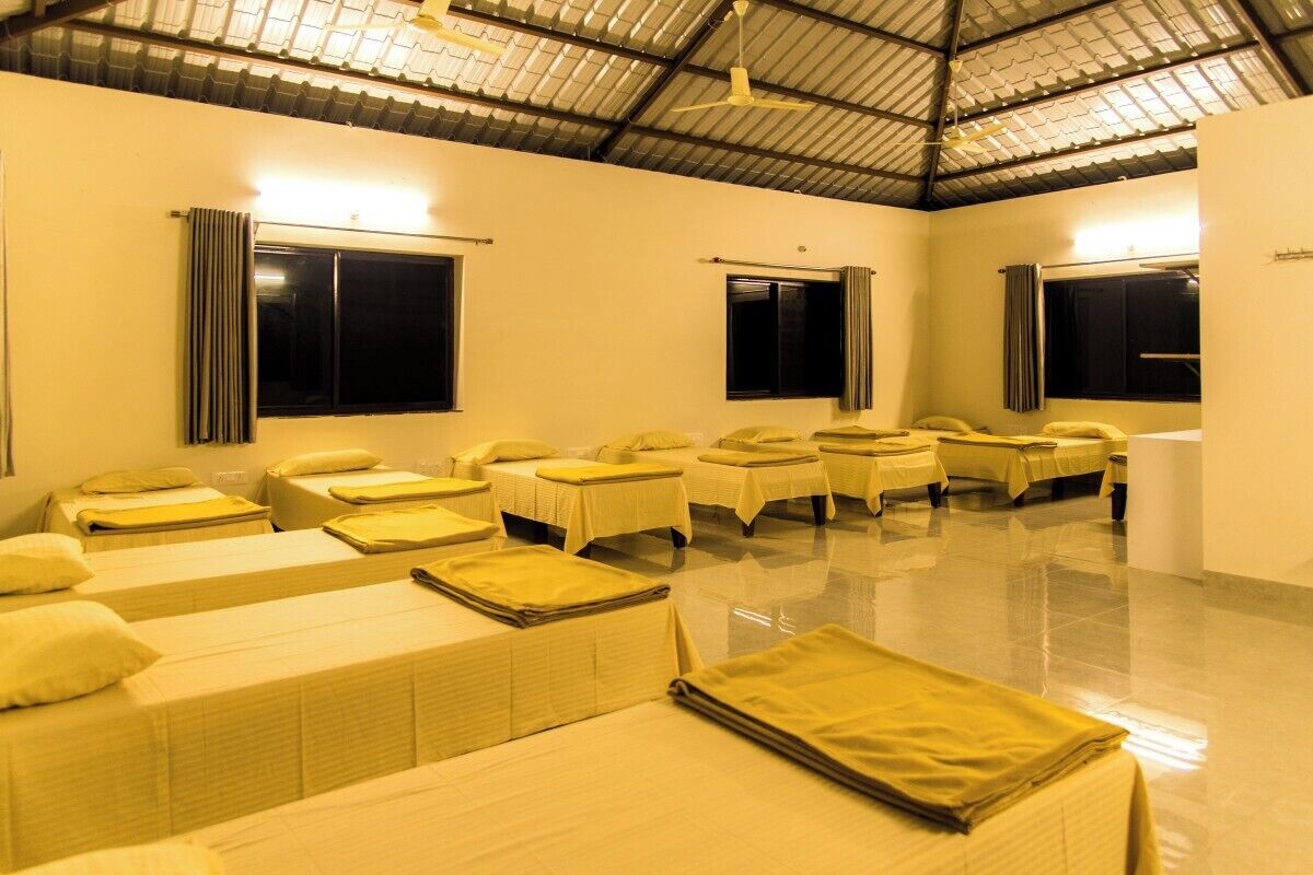 A room with multiple beds arranged in rows, a high ceiling, and large windows with curtains.