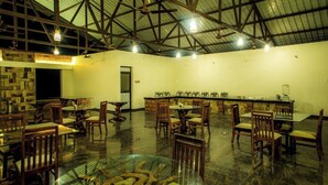 A spacious indoor area with multiple dining tables and chairs, a stone fireplace, and a high ceiling with metal beams. - Parampara Cottages (Haliyal)