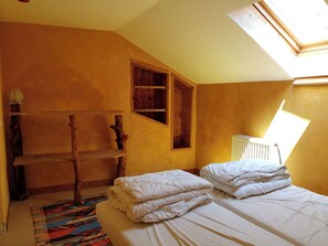 A room with a sloped ceiling, a skylight, a bed with white bedding, a wooden shelf, and a colorful rug.
