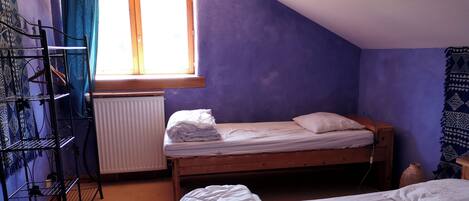 Ein kleines Zimmer mit einem Einzelbett, geneigter Decke, Heizkörper, einem mit Vorhängen versehenen Fenster, einem Wäscheständer aus Metall und einem gemusterten Teppich.