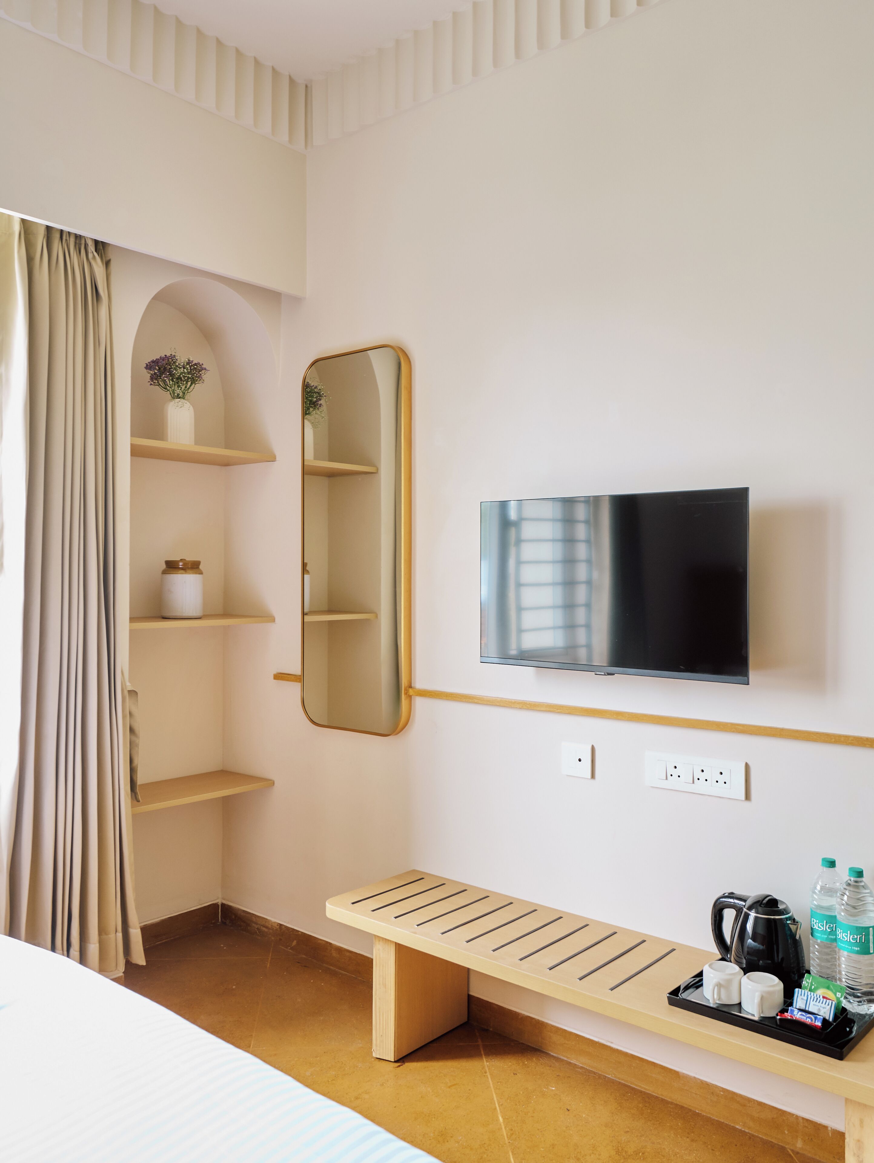 A hotel room with a bed, a TV mounted on the wall, a wooden bench, a shelf with a vase, and a window with curtains.