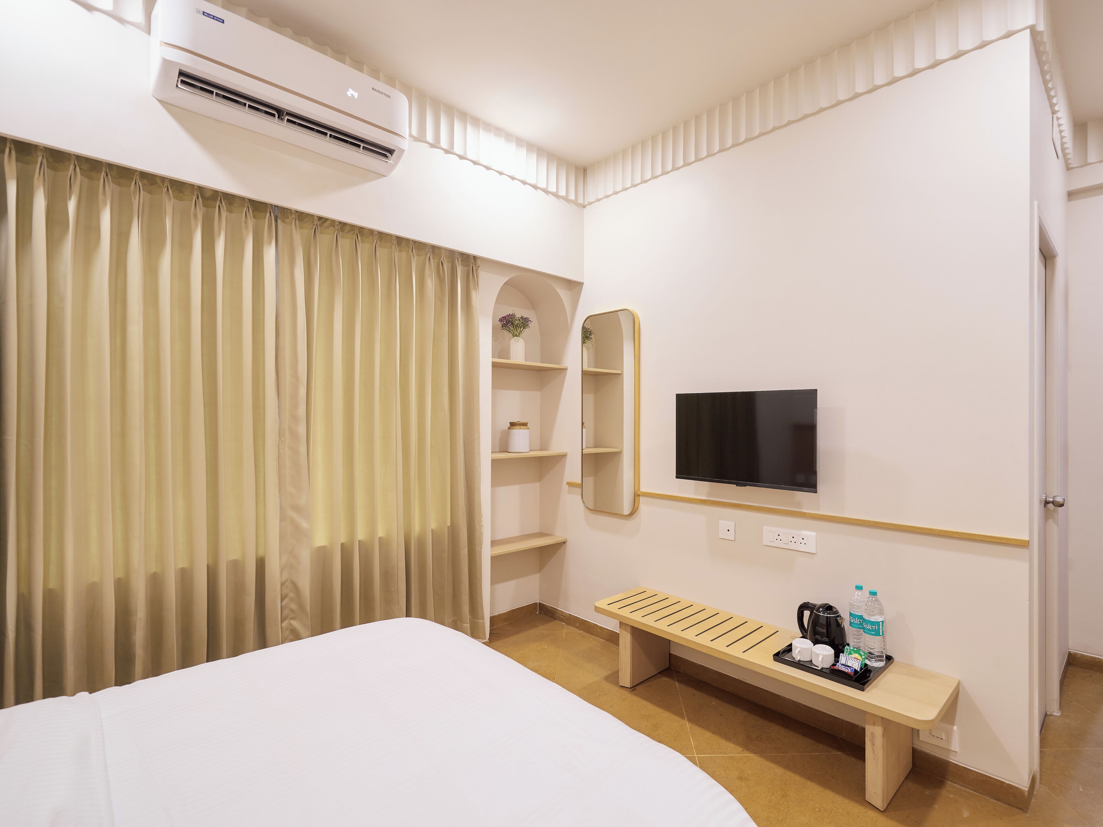 A hotel room with a bed, an air conditioning unit, a TV, a mirror, a shelf, and a bench with amenities.