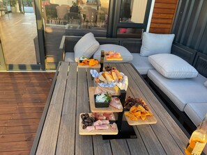 Een houten terras met een glazen zithoek, inclusief een bank en een tafel met verschillende soorten eten.