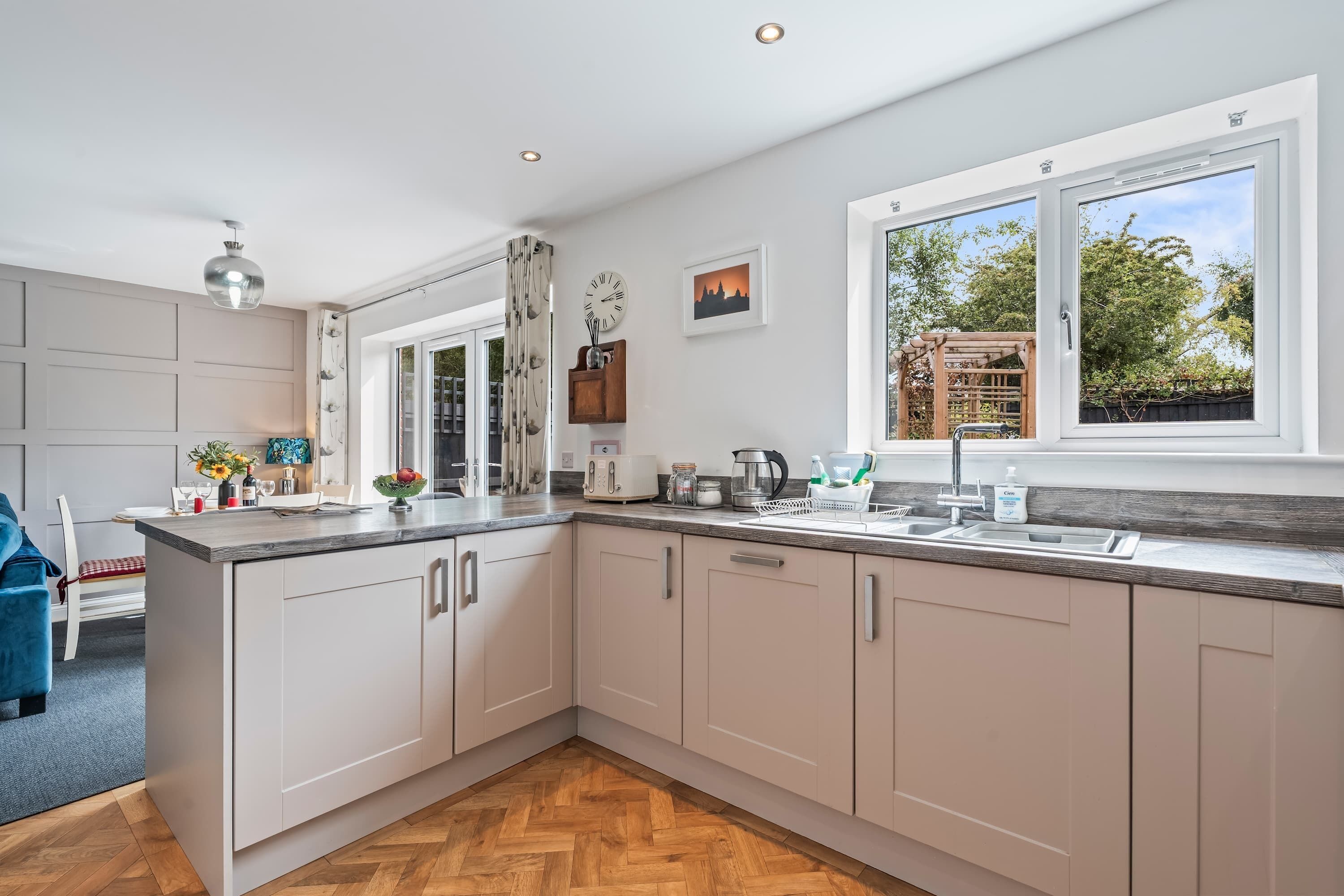 Une cuisine moderne avec un parquet en bois disposé en chevrons, des meubles blancs, un îlot central intégré et une fenêtre donnant sur l’extérieur.