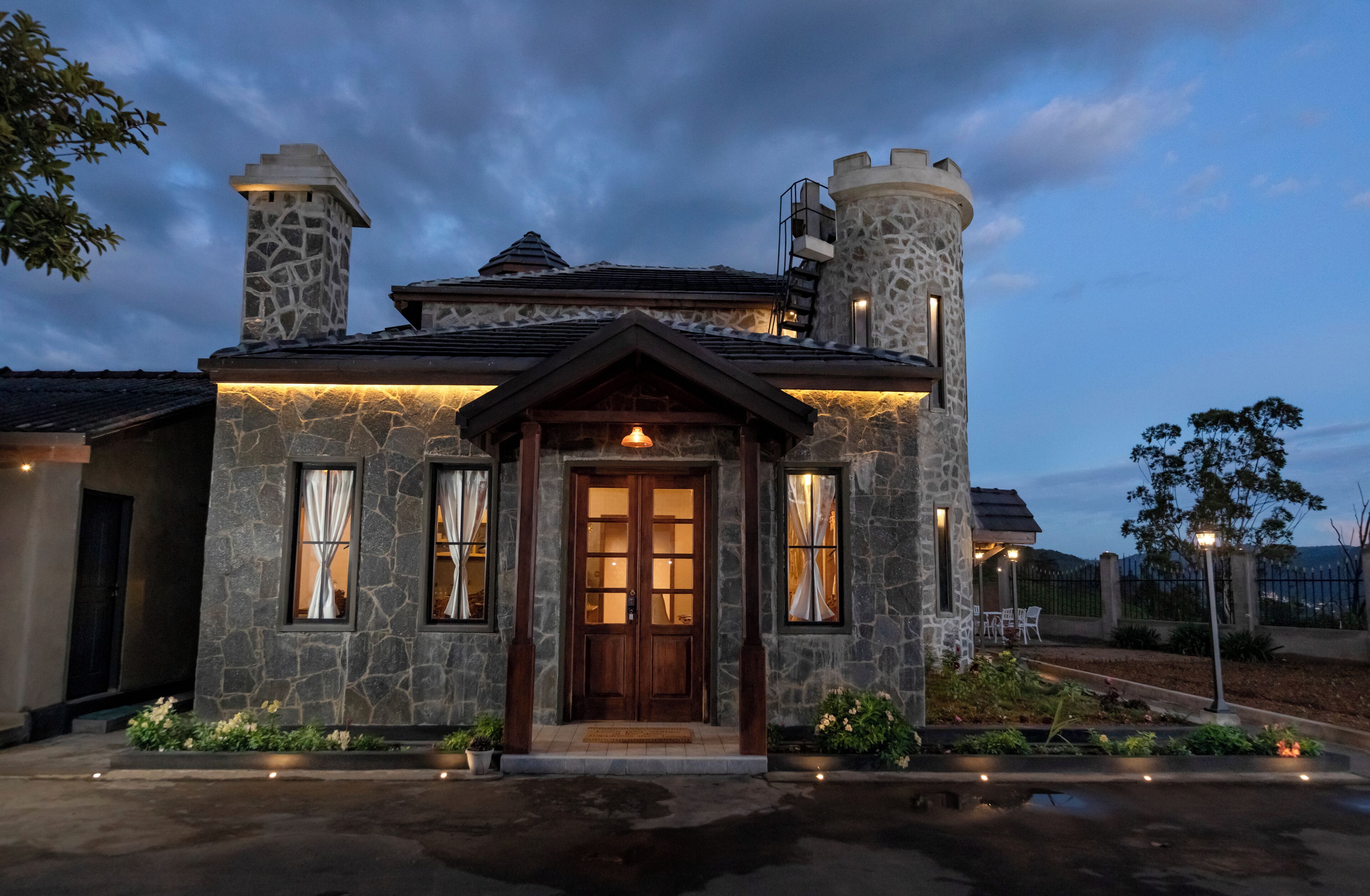 Uma casa de pedra com porta de madeira, duas chaminés e um pequeno alpendre com cadeiras.