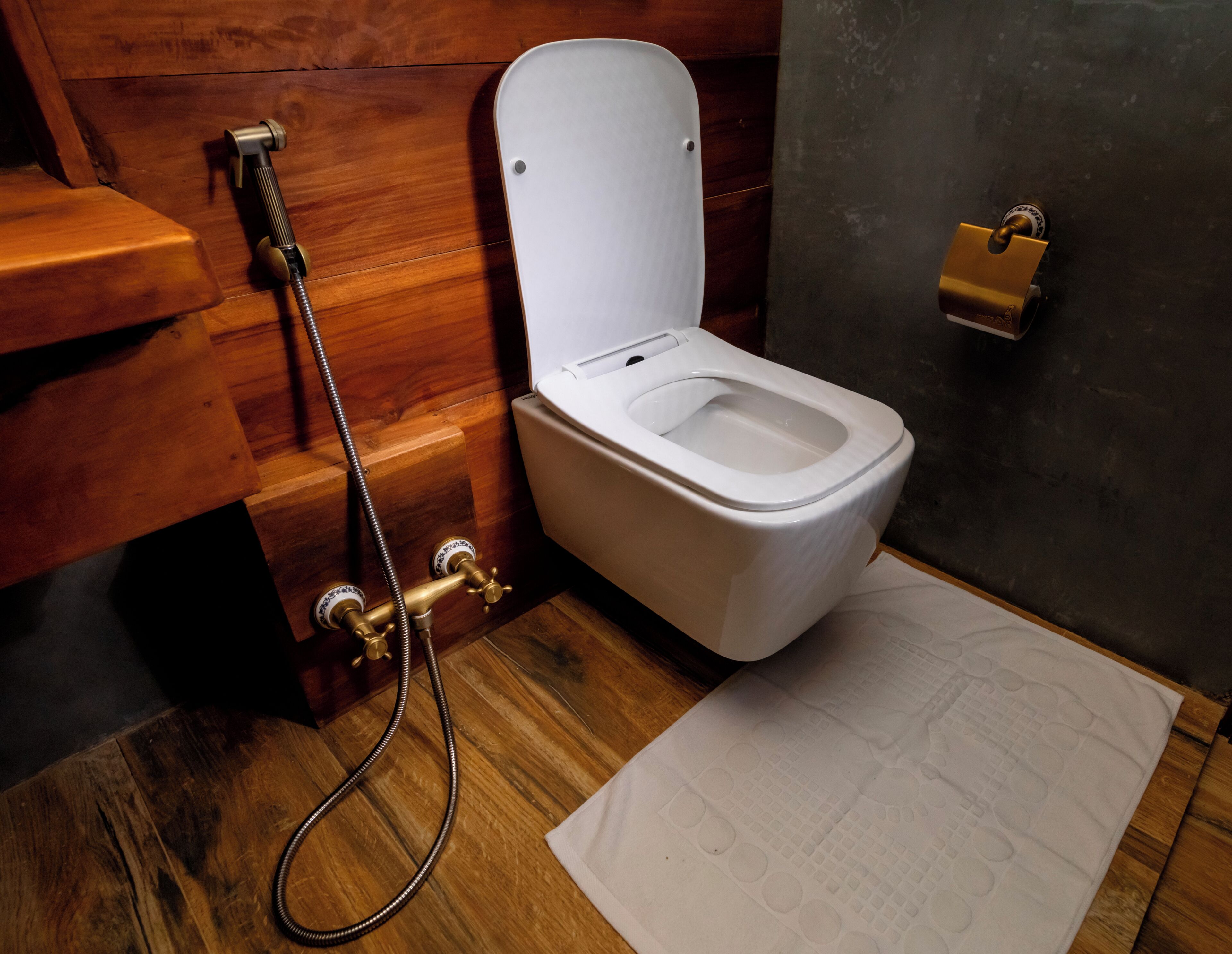 A wooden toilet with a bidet attachment, a paper towel holder, and a wooden floor.