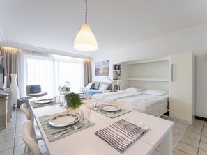 Una habitació d'hotel moderna amb zona de menjador, llit, armari i una finestra gran amb cortines.