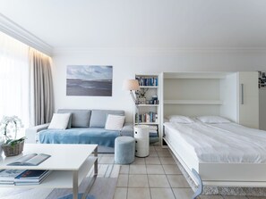 Une chambre d’hôtel moderne avec un canapé, un lit, une bibliothèque et un tableau représentant une plage.