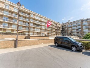 Un immeuble résidentiel à plusieurs étages, doté de balcons, d’un espace de stationnement et d’un fourgon noir garé devant.