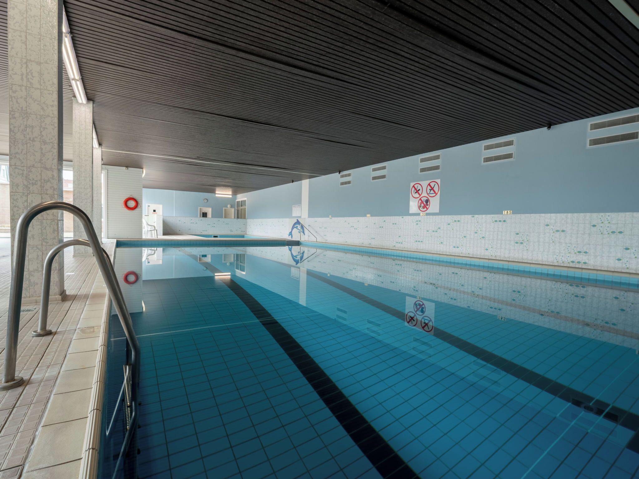 An indoor swimming pool with tiled walls, a clear water surface, and a ceiling with a ribbed pattern.