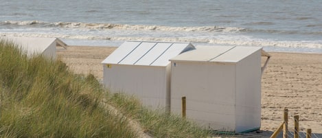 Una playa con un sistema de dunas, un pequeño edificio blanco y un velero a lo lejos.