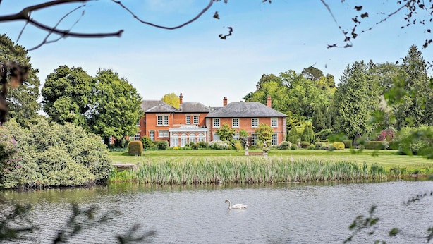 Een groot stenen huis met een vijver op de voorgrond en een verzorgde tuin.