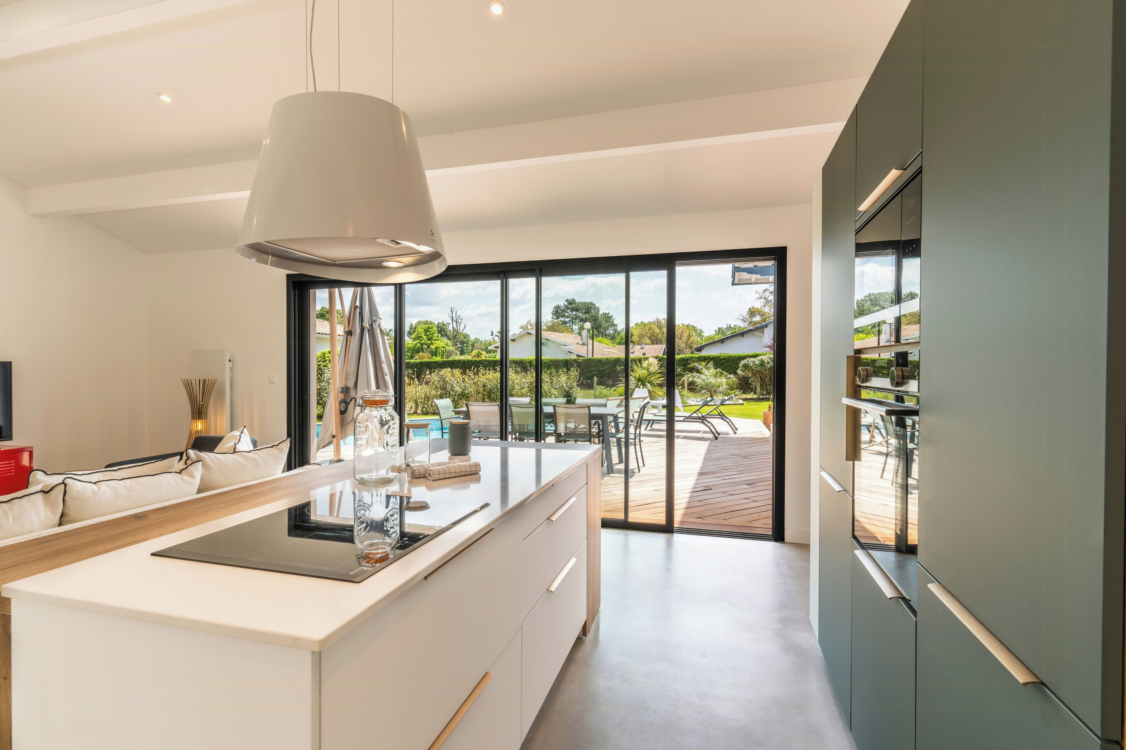 A modern kitchen with a central island, a dining area with outdoor seating, and a view of a garden and pool.