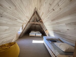 Um quarto triangular com paredes de madeira e teto inclinado, contendo duas camas e um pufe amarelo.