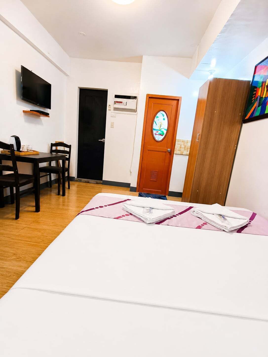 A hotel room with a bed, a desk with chairs, a television, and a door with a clock.