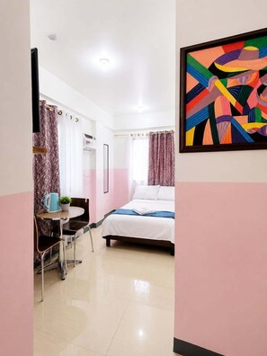 A hotel room with a bed, a small table with a kettle and a plant, a chair, a window with curtains, and a colorful abstract painting on the wall.