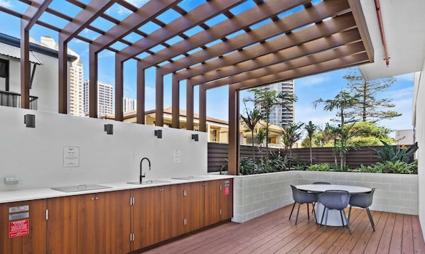 An outdoor kitchen area with a built-in sink, wooden cabinets, and a pergola overhead. - Opus 2 Bedroom Ocean (Broadbeach)
