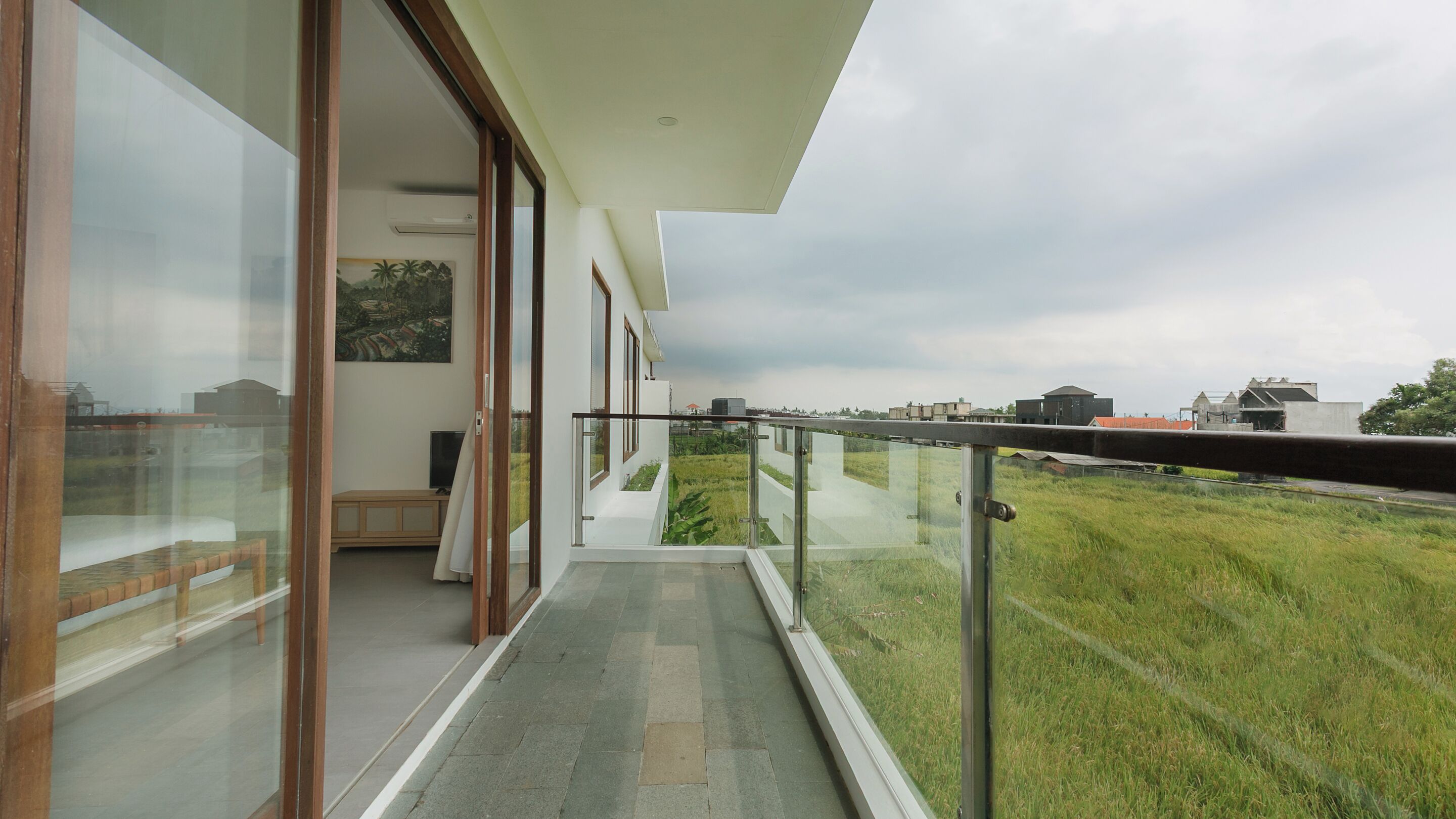 A modern balcony with a glass railing overlooking a grassy area and distant buildings.