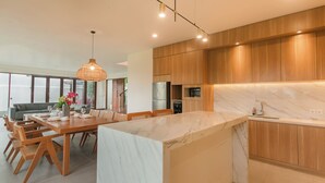 A modern kitchen with wooden cabinets, a marble countertop, and a dining area with a table and chairs. - Ombak D3 Villa by Betterplace (Cemagi)