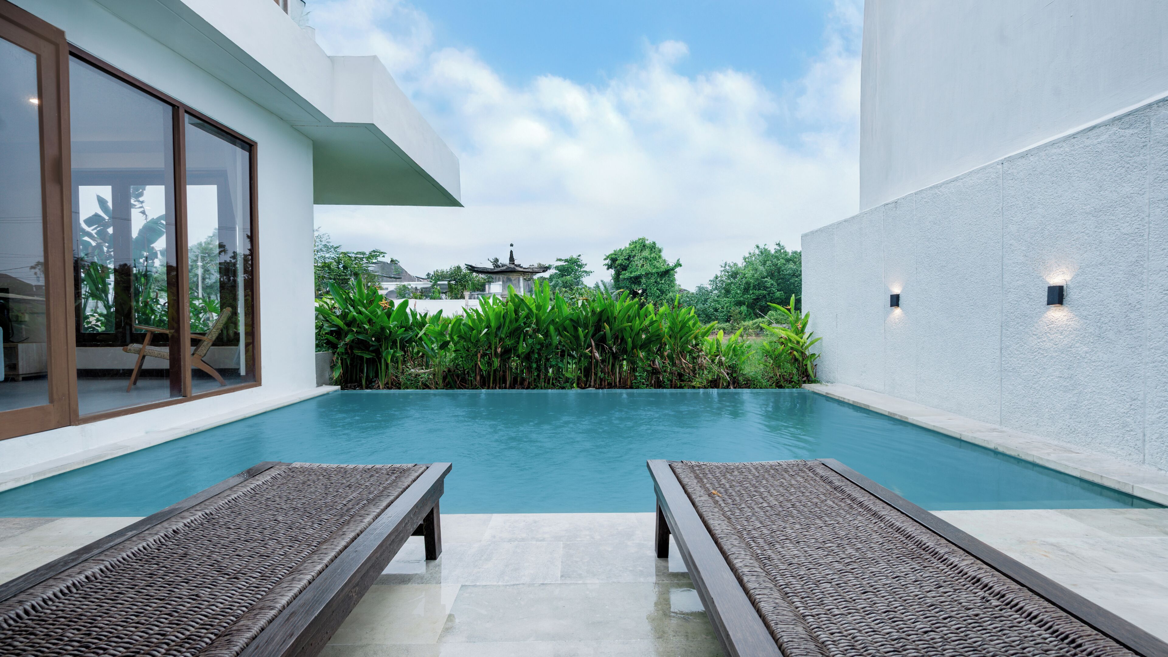 Uma área de piscina moderna com duas espreguiçadeiras, uma porta de vidro que dá para um quarto, e vista para um edifício tradicional ao fundo.