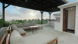 A covered outdoor seating area with a sofa and a table, a door leading to a building, and a view of greenery and a distant hill. - Ombak D3 Villa by Betterplace (Cemagi)
