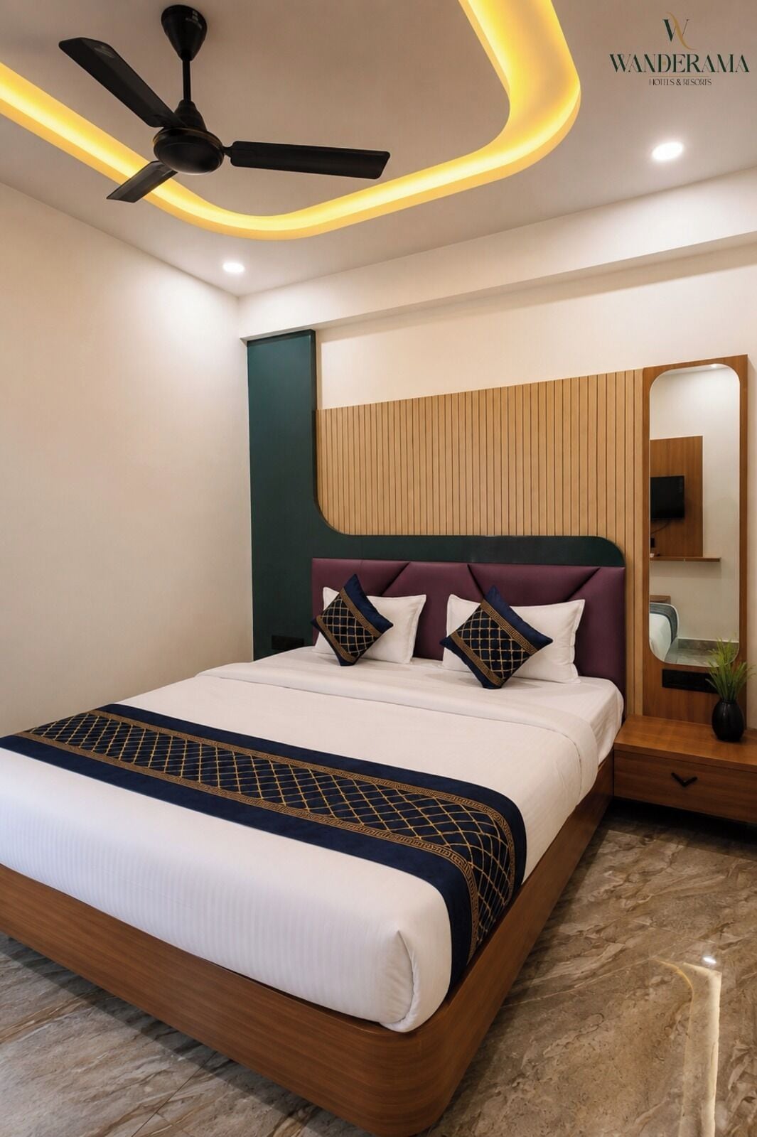 Une chambre d’hôtel avec un lit, un ventilateur de plafond, une tête de lit en bois, une table de chevet, un miroir et une plante.
