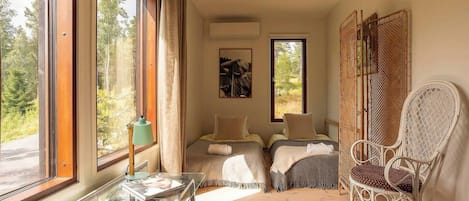 Une chambre avec deux lits, un fauteuil en osier, une table avec une lampe et de grandes fenêtres avec des rideaux.