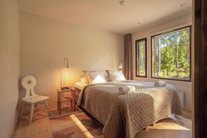 A bedroom with a large bed, a bedside table with a lamp, a chair, and a view of trees through the window.