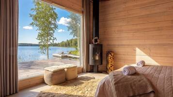 Een kamer met een groot raam met uitzicht op een meer, een open haard, een bed met opgerolde handdoeken en een houten vloer.