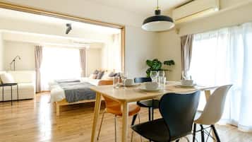 A well-lit hotel room with a wooden floor, a bed with a white headboard, a dining table set with plates and glasses, and a potted plant.