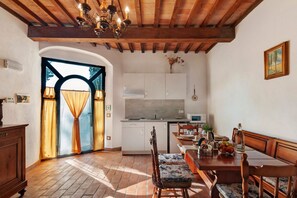 A dining area with a wooden table, chairs, and a chandelier. There is a kitchen in the background with cabinets and a microwave. - Rosmarinus Apartment – Two-Room Flat with Views of Tuscan Vineyards (San Casciano In Val di Pesa)
