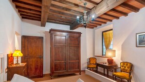 A room with a bed, a wooden wardrobe, a table with chairs, a chandelier, and a window with curtains. - Rosmarinus Apartment – Two-Room Flat with Views of Tuscan Vineyards (San Casciano In Val di Pesa)