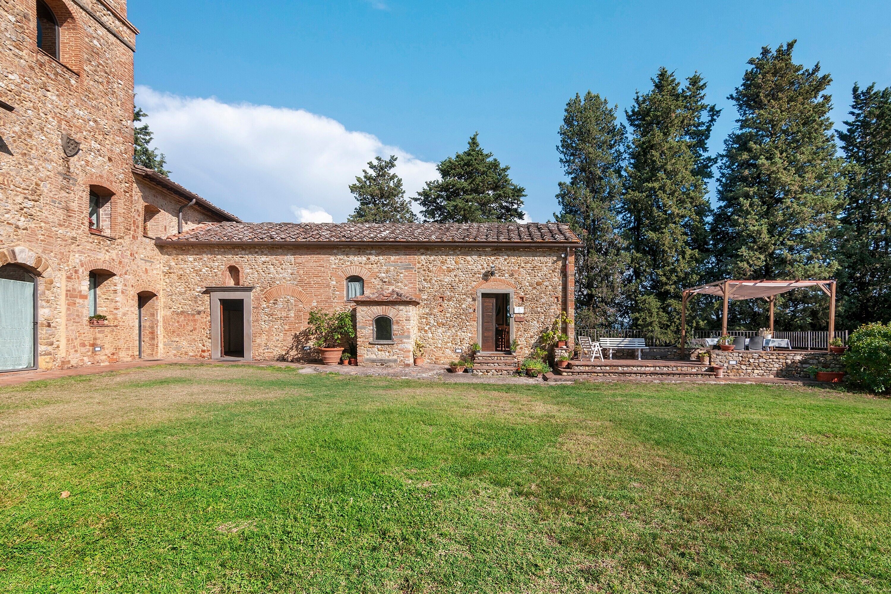 Un edificio de piedra con techo de tejas, un patio cubierto de césped y una pérgola de madera con asientos.