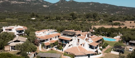 A residential area with houses featuring terracotta roofs, a swimming pool, and a mountainous backdrop.