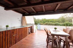 An outdoor patio with a wooden table and chairs, a built-in grill, and a view of greenery. - Vera Holiday Home by Wonderful Italy (Olbia)