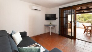 Una sala de estar con un sofá, una mesita con una planta, un televisor de pantalla plana y un ventanal corredero que da a un espacio exterior con una mesa y sillas.