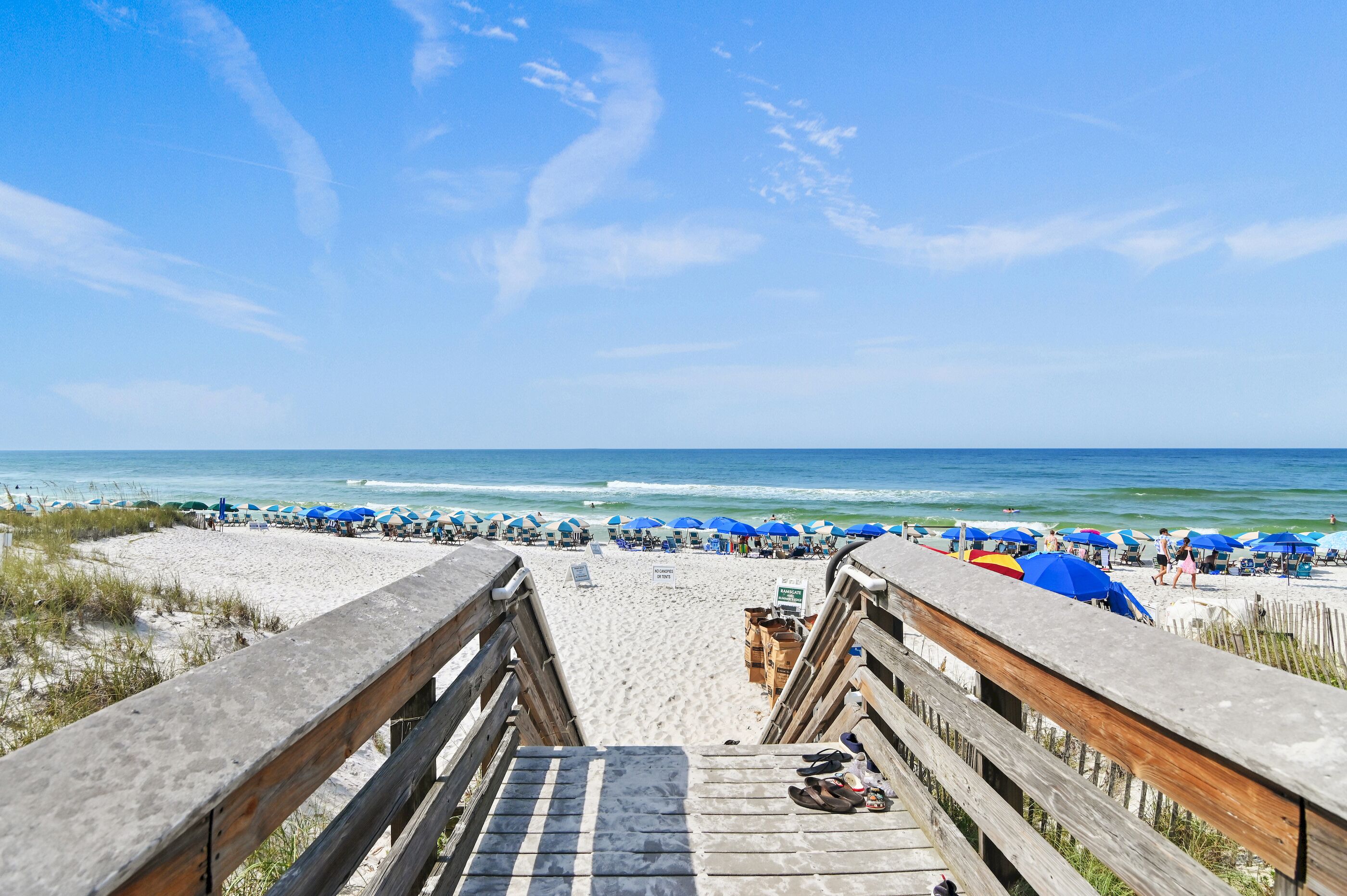 A wooden boardwalk leading to a beach with rows of blue umbrellas and beach chairs.